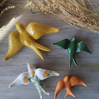 Quatro estatuetas de pássaros coloridos em cerâmica sobre mesa de madeira clara com ramo seco