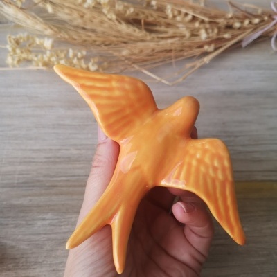 Pássaro decorativo de cerâmica laranja segurado por uma mão com ramos secos ao fundo