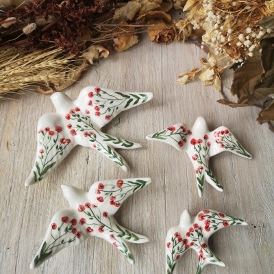 Figuras de pássaros em cerâmica branca pintadas com flores vermelhas e folhas verdes em madeira com flores secas.