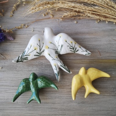 Figuras decorativas de pássaros de cerâmica em branco com flores, verde e amarelo sobre mesa de madeira com trigo seco