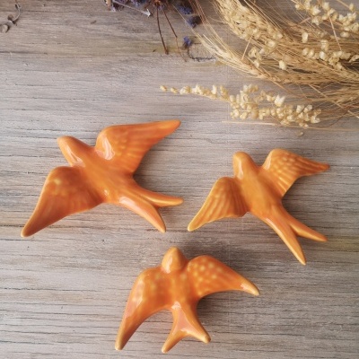 Três figuras decorativas em cerâmica de pássaros alaranjados sobre mesa de madeira com ramos secos.