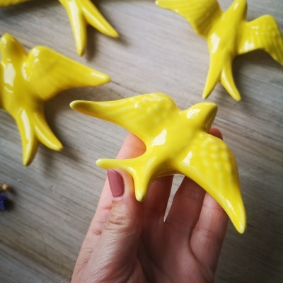 Figuras de pássaros amarelos em cerâmica com textura, sobre madeira e mão a segurá-los.