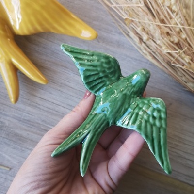 Estatueta cerâmica verde em forma de pássaro com asas abertas, mão segurando, ramos secos e superfície de madeira