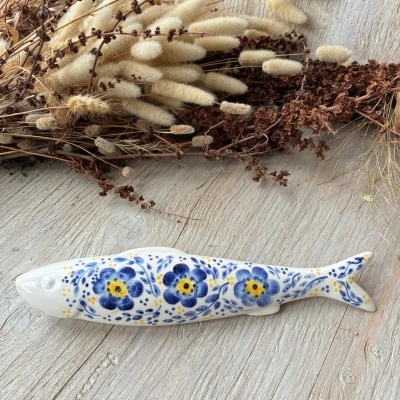 Peixe decorativo em cerâmica com padrão de flores azuis e amarelas sobre superfície de madeira clara.