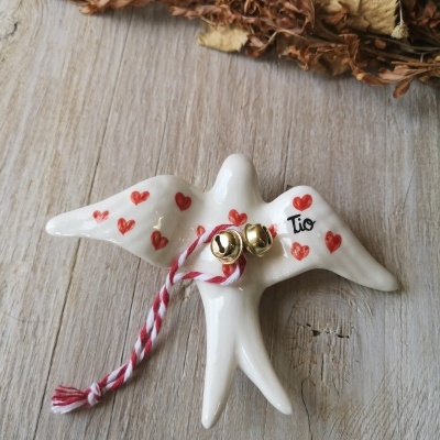 Figurinha branca de pássaro com corações vermelhos e texto Tio, com cordão e guizos dourados, em madeira clara.