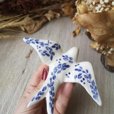 Escultura de pássaro em cerâmica branca com folhas azuis, mão segurando, fundo madeira e folhas secas