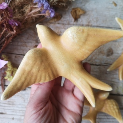 Escultura de pássaro em madeira clara, textura visível, mão segurando, flores secas ao fundo