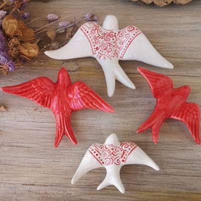 Figuras decorativas de pássaros em cerâmica branca e vermelha sobre madeira clara com flores secas.