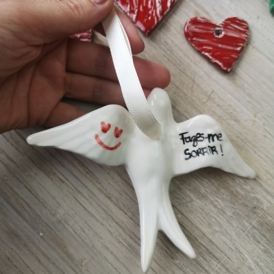 Pássaro de cerâmica branco com fita, texto e rosto sorridente, sobre mesa com corações vermelhos