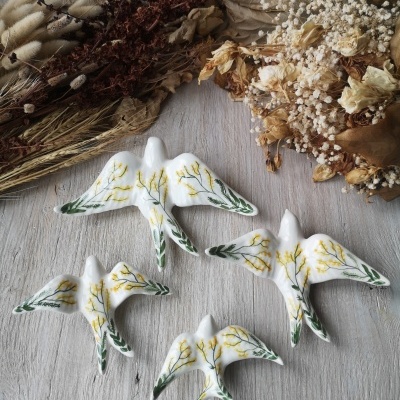 Figuras de cerâmica branca com padrão floral em forma de pássaros sobre madeira clara com flores secas ao fundo