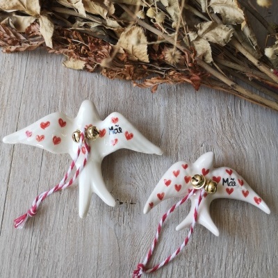 Pássaros decorativos brancos com corações vermelhos e texto Mãe, sinos dourados e fitas vermelhas e brancas, sobre mesa de madeira clara e folhas secas.