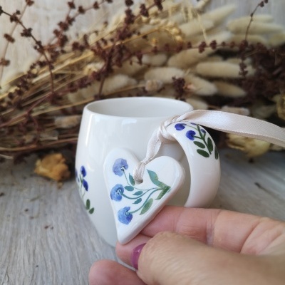 Caneca branca com padrão floral azul e verde, pingente coração e fundo de flores secas.