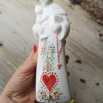 Estátua religiosa branca com padrões coloridos e terço vermelho, em fundo de madeira