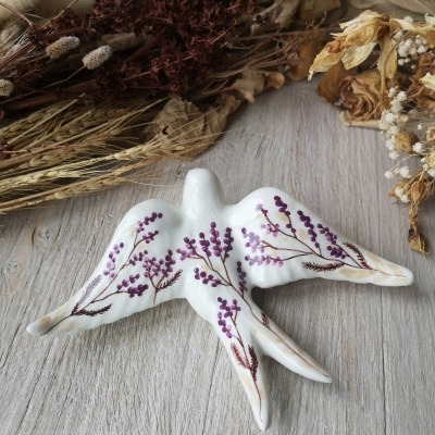 Peça decorativa de cerâmica branca em forma de pássaro com pintura floral roxa sobre superfície de madeira clara.