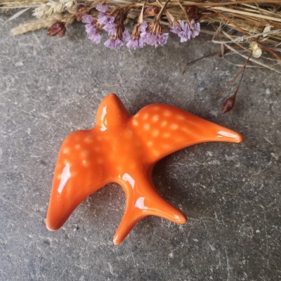 Peça decorativa de cerâmica em forma de pássaro laranja sobre superfície cinza com flores secas