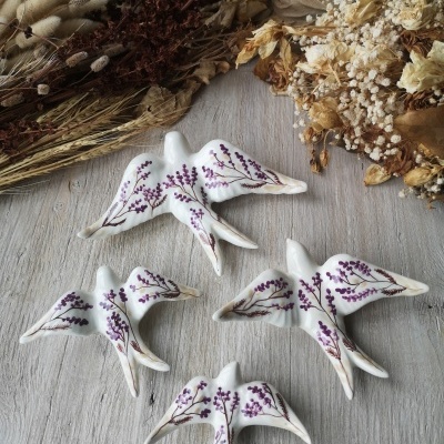 Pássaros decorativos em cerâmica branca com flores roxas, sobre mesa de madeira, com flores secas ao redor