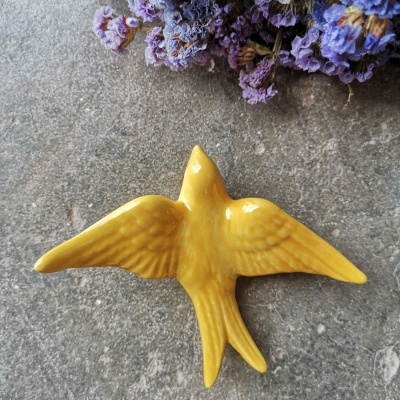 Pássaro decorativo em cerâmica amarela sobre superfície cinza com flores secas roxas.