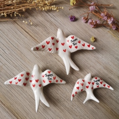 Três andorinhas cerâmicas brancas com corações vermelhos e texto 'Super Tia' sobre mesa de madeira com flores secas
