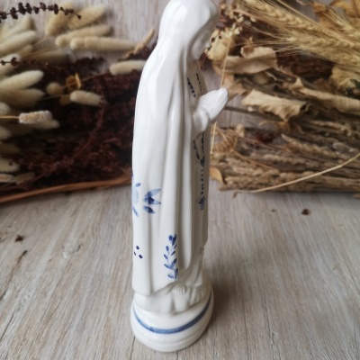 Estátua branca de cerâmica de Nossa Senhora com detalhes azuis e fundo de plantas secas