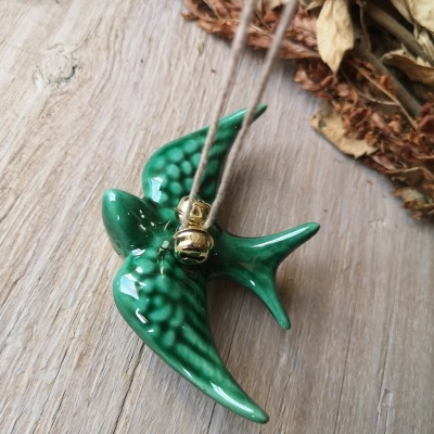 Pendente cerâmico verde em forma de andorinha com sino dourado sobre madeira clara