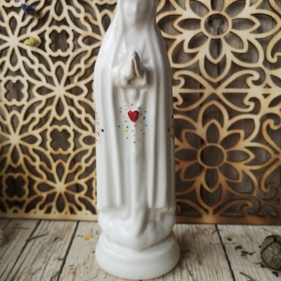Estátua branca de porcelana com figura religiosa e coração vermelho em fundo de padrão floral de madeira