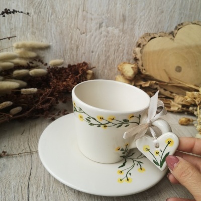 Conjunto de chávena e pires com padrão floral amarelo e verde, com coração decorativo e laço bege.