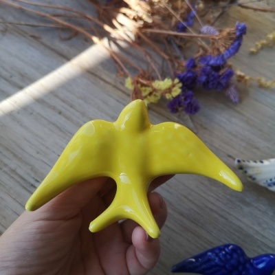Adorno cerâmico amarelo em forma de pássaro na mão, com flores secas e pássaro azul ao fundo