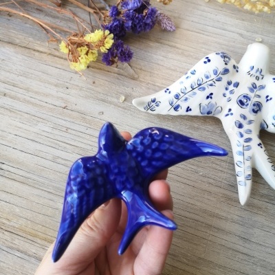 Figura decorativa de pássaros em cerâmica azul e branca com flores secas ao fundo sobre mesa de madeira