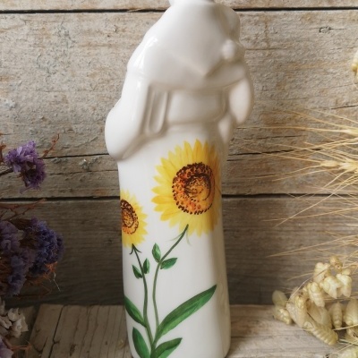 Figura decorativa branca em cerâmica com pintura de girassóis, sobre fundo de madeira rústica e flores secas
