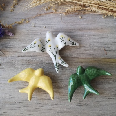 Três pássaros em cerâmica coloridos sobre madeira clara com trigo seco ao fundo