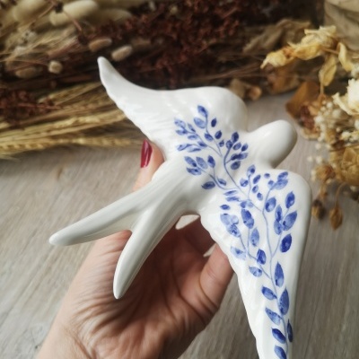 Escultura branca de pássaro com pintura azul em forma de folhas, segurada por uma mão, sobre fundo de madeira clara com flores secas