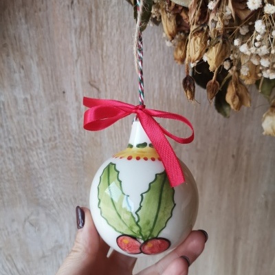 Bola de Natal cerâmica branca com folhas verdes e bagas vermelhas, laço vermelho, mão segurando, fundo madeira e flores secas