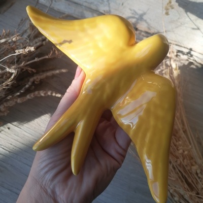 Pássaro decorativo amarelo em cerâmica segurado à mão, sobre fundo de madeira clara