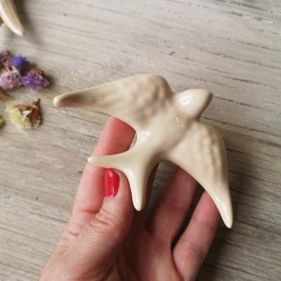 Figura de pássaro em cerâmica creme sendo segurada por mão com unhas vermelhas sobre fundo de madeira clara com flores secas.