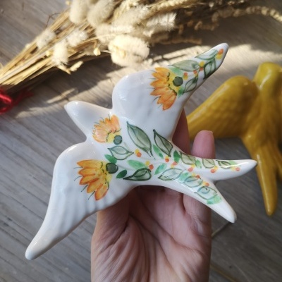 Pássaro decorativo em cerâmica pintada com flores e folhas, colocado sobre fundo de madeira com ramos secos