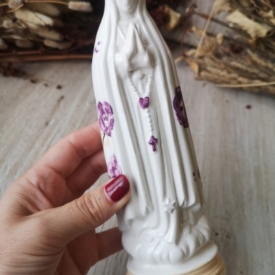 Estatueta religiosa de porcelana branca com flores roxas e terço roxo, segura por mão com unhas vermelhas