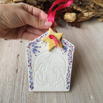Ornamento de cerâmica branca com relevo de Natividade e estrela amarela com fita vermelha pendurada