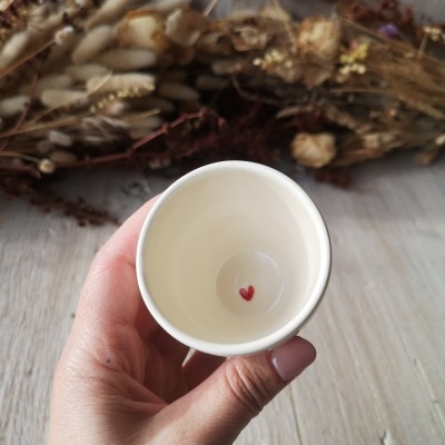 Copo de cerâmica creme com coração vermelho na base, sendo segurado por mão feminina sobre mesa de madeira clara.