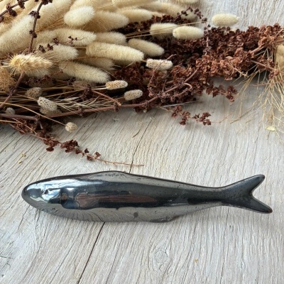 Peixe decorativo de metal preto brilhante ao lado de flores secas castanhas e beiges sobre madeira clara.