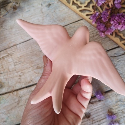 Escultura de pássaro rosa claro em cerâmica segurada por mão, com fundo de madeira e flores secas roxas