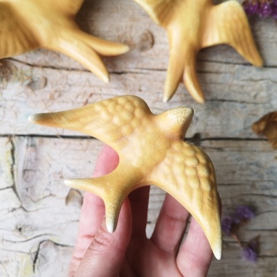 Figuras decorativas de pássaros amarelo-alaranjados com textura, sobre madeira clara, uma mão segura um deles