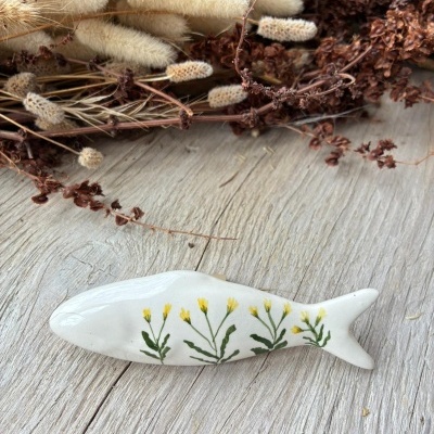 Peixe decorativo em cerâmica branca com flores amarelas pintadas e folhas verdes sobre madeira clara