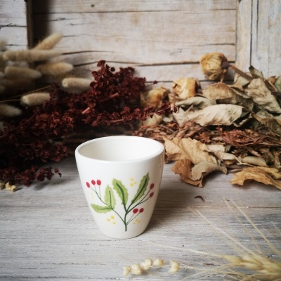 Copo branco de cerâmica com desenho de folhas e frutos vermelhos sobre madeira com ramos secos
