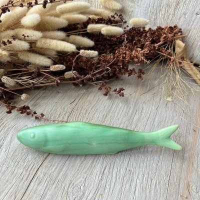 Peixe decorativo em cerâmica verde claro próximo a plantas secas em mesa de madeira.