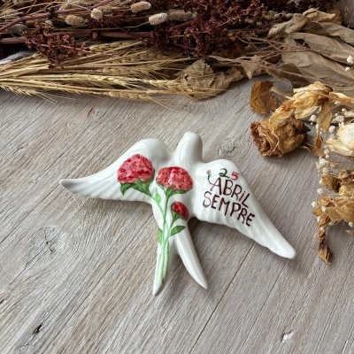 Estatueta de cerâmica branca em forma de pomba com flores vermelhas e texto 25 ABRIL SEMPRE sobre mesa de madeira clara