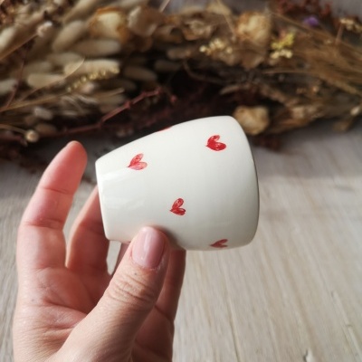 Copo de cerâmica branca com corações vermelhos segurado por mão em fundo de madeira clara e flores secas.