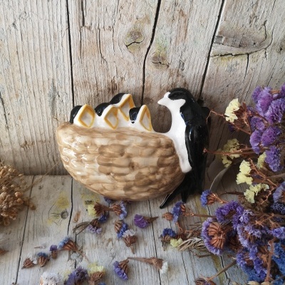 Figura decorativa de cerâmica com pássaro e pintainhos numa cesta, sobre madeira com flores secas.