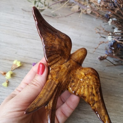 Estatueta cerâmica de pássaro castanho brilhante numa mão com unhas vermelhas e fundo de madeira com flores secas