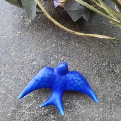 Pássaro azul decorativo em cerâmica ou resina sobre superfície cinza com folhas e flores secas