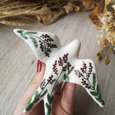 Pássaro decorativo branco com flores pintadas e mão a segurá-lo sobre fundo de madeira e flores secas